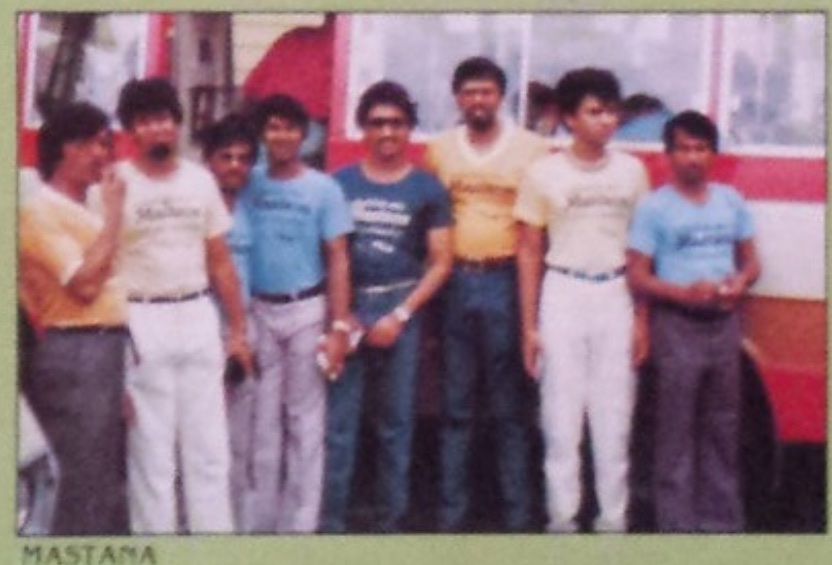 Faded colour photo of the Mastana Orchestra, eight musicians standing in a row in front of a tour bus