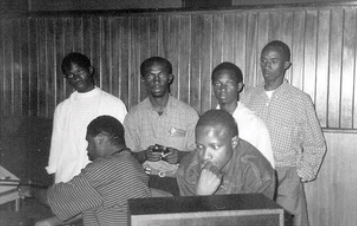 Black-and-white photo of six young men gathered around a mixing desk in a wood-panelled studio